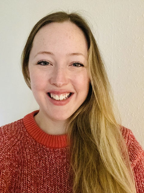 DR with long mid brown hair wearing a red jumper against a cream background